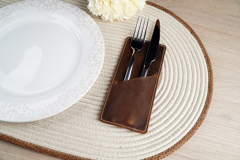 May include: A brown leather cutlery holder with a fork and knife inside. The holder is on a white and brown woven placemat.