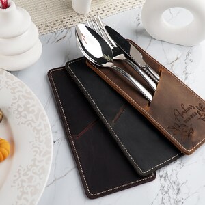 May include: A brown leather cutlery roll with the name "Amber's Kitchen" engraved on it. The roll is open and contains a fork, knife, and spoon.