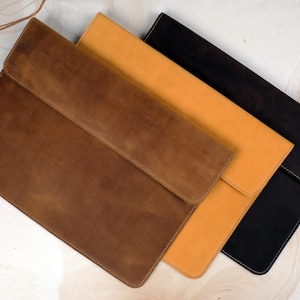 May include: Three rectangular document sleeves with fold-over flaps, arranged in a diagonal stack. The sleeves are colored in dark brown, tan, and black, each featuring visible stitching along the edges and a smooth, matte surface texture.
