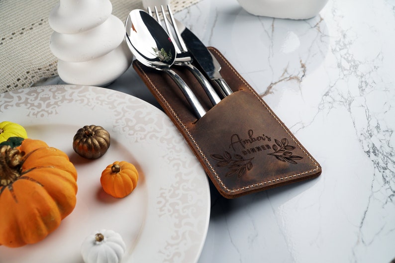 May include: A brown leather cutlery holder with the text "Amber's Dinner" engraved on it. The holder has a pocket for a spoon, fork, and knife.