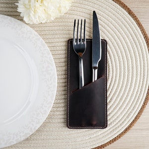 May include: A brown leather cutlery holder with a fork and knife inside. The holder is on a white woven placemat.