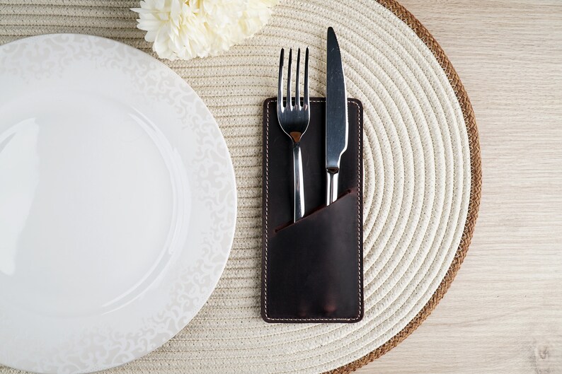 May include: A brown leather cutlery holder with a fork and knife inside. The holder is on a white woven placemat.