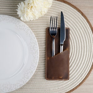 May include: A brown leather cutlery holder with a silver fork and knife inside. The holder is on a white round placemat with a white plate next to it.