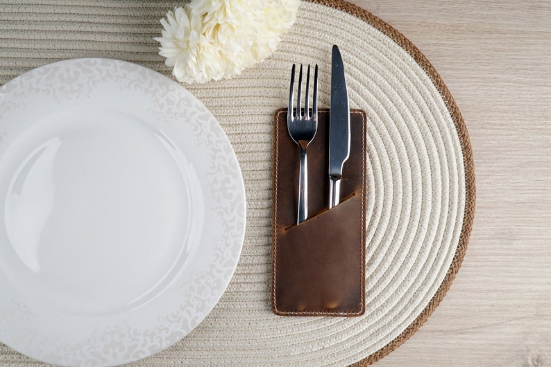 May include: A brown leather cutlery holder with a silver fork and knife inside. The holder is on a white round placemat with a white plate next to it.