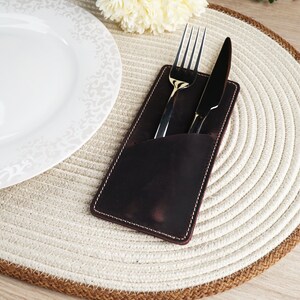May include: A brown leather cutlery holder with a white stitch detail, holding a silver fork and knife on a white and brown woven placemat.