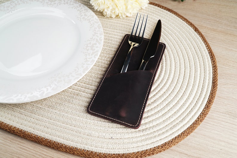 May include: A brown leather cutlery holder with a white stitch detail, holding a silver fork and knife on a white and brown woven placemat.