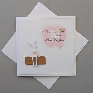 May include: A white greeting card with a pink watercolour splash and the text "Willkommen kleine Mia Sophie." A wooden bunny figure sits on a wooden base. A small wooden star is also on the card. The card is inside a white envelope.