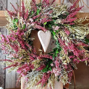 Könnte beinhalten: Ein dekorativer Kranz mit einem Holzherz in der Mitte, umgeben von einer Mischung aus getrockneten Blumen und Grün. Der Kranz enthält rosa Heidekraut, beige Gräser und grünes Laub, was eine rustikale und natürliche Ästhetik schafft.