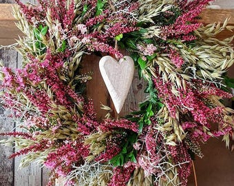 May include: A decorative wreath featuring a wooden heart in the center, surrounded by a mix of dried flowers and greenery. The wreath includes pink heather, beige grasses, and green foliage, creating a rustic and natural aesthetic. The background is a wooden surface.