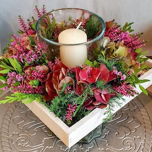 Könnte beinhalten: Ein quadratisches Tischgesteck in einer weißen Holzbox mit einem klaren Glas-Kerzenhalter und einer cremefarbenen Stumpenkerze, umgeben von rosa Heidekraut, dunkelroten Hortensienblüten und grünem Nadelgehölz in dichter Anordnung.