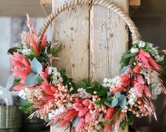 May include: A circular wreath made of a rope base, adorned with pink, white, and green dried flowers and foliage. The wreath is suspended against a rustic wooden background, creating a natural and decorative accent.