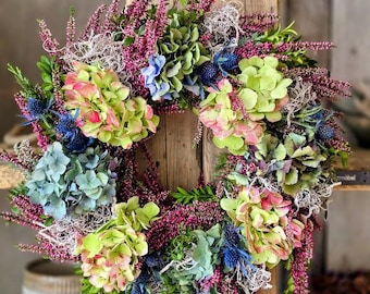 Op de afbeelding: Een ronde krans samengesteld uit felroze heide, lichtgroene en blauwe hortensiabloesems en stekelige blauwe distels. Het arrangement is afgewisseld met groen blad en ijle, lichtgekleurde gedroogde mos, wat zorgt voor een dicht en textuurrijk bloemstuk.