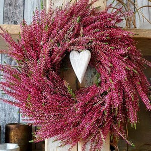 Könnte beinhalten: Ein runder Kranz aus leuchtend magentafarbenen Heidekrautblüten mit einem mittig hängenden, weißen Holzherz an einer dünnen Schnur. Das dichte, strukturierte Blumenarrangement bildet eine volle, runde Form mit sichtbaren grünen Stielen.