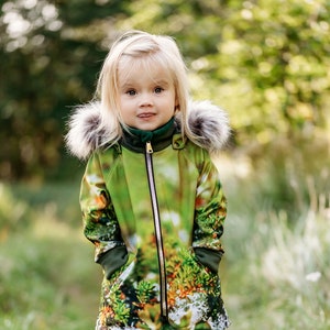May include: A young girl wearing a green and brown patterned jacket with a fur-trimmed hood. She is standing on a path in a forest.
