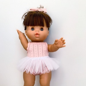 May include: A brown-haired doll wearing a pink and white dress with a white tulle skirt. The doll has brown eyes and is wearing a pink bow in her hair.