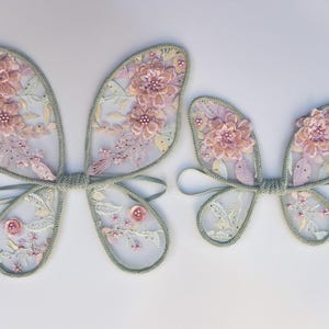 May include: Two sets of fairy wings. The wings are made of sheer, light-colored fabric with a floral design in shades of pink and white. The edges are trimmed with a light green trim. The wings are connected by a light green tie.