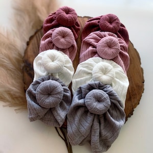 Pode incluir: Uma coleção de turbantes para bebés em várias cores, incluindo malva, branco e cinza. Cada turbante apresenta um detalhe de nó na parte superior. O tecido parece ser canelado, adicionando textura. Os turbantes estão dispostos sobre uma superfície de madeira.