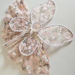 May include: A pair of sheer butterfly wings featuring intricate floral embroidery in shades of pink, cream, and sage green, accented with shimmering sequins. The wings are outlined with a silver-toned trim and include thin white ribbons for attachment.