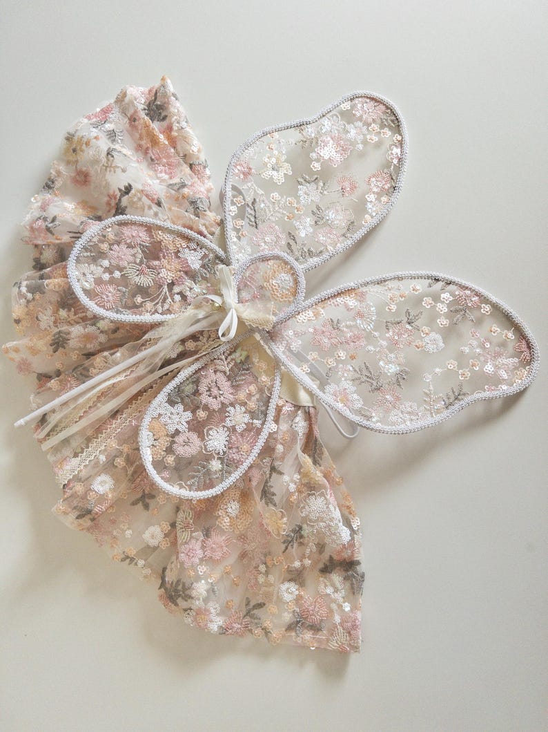 May include: A pair of sheer butterfly wings featuring intricate floral embroidery in shades of pink, cream, and sage green, accented with shimmering sequins. The wings are outlined with a silver-toned trim and include thin white ribbons for attachment.