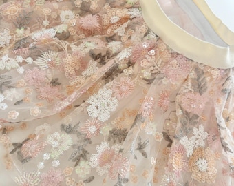 May include: A delicate tulle skirt with a floral pattern and sequins. The skirt is a light pink color with a cream-colored waistband.