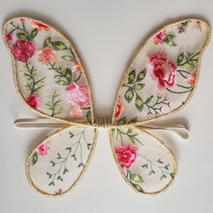 May include: Delicate butterfly wings with a sheer, cream-colored fabric, adorned with embroidered pink and red flowers and green leaves. The edges are trimmed with gold-colored thread. A white elastic band is attached for wear.