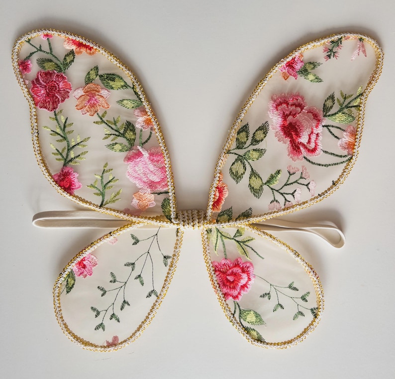 May include: Delicate butterfly wings with a sheer, cream-colored fabric, adorned with embroidered pink and red flowers and green leaves. The edges are trimmed with gold-colored thread. A white elastic band is attached for wear.