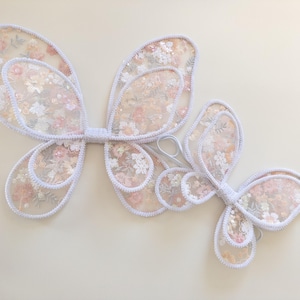 May include: A pair of butterfly wings and a smaller single butterfly wing accessory, both crafted from sheer white fabric with a delicate floral pattern in shades of pink, peach, and green, featuring white stitched edges and elastic loops.