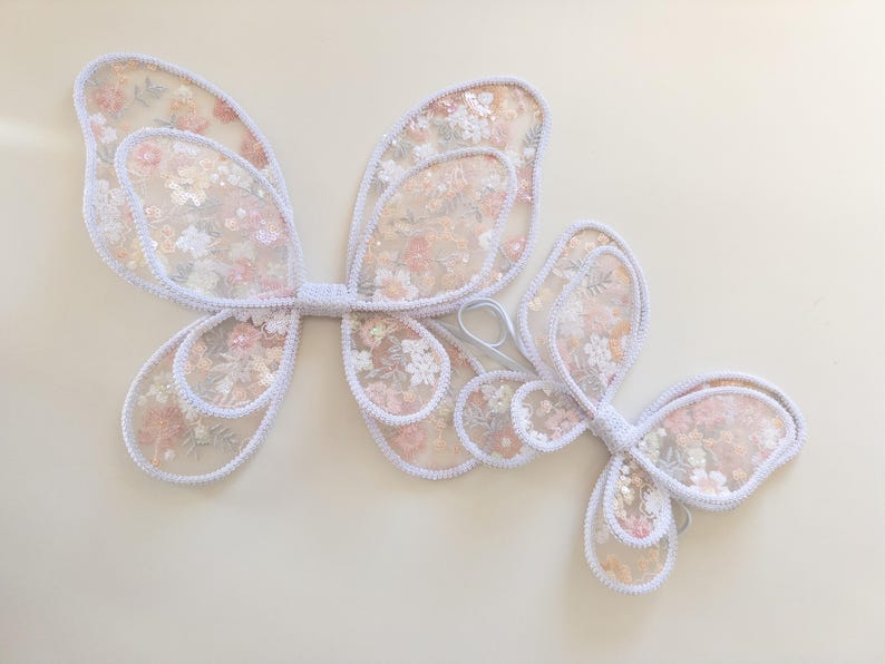 May include: A pair of butterfly wings and a smaller single butterfly wing accessory, both crafted from sheer white fabric with a delicate floral pattern in shades of pink, peach, and green, featuring white stitched edges and elastic loops.