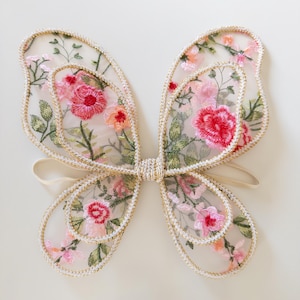 May include: Delicate butterfly wings with a floral design. The wings feature a sheer, cream-colored fabric embroidered with pink, red, and orange flowers, outlined with gold trim. A cream-colored elastic band connects the wings.