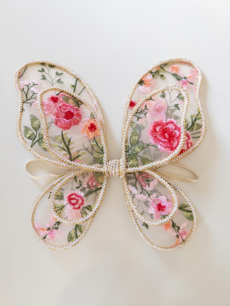 May include: Delicate butterfly wings with a floral design. The wings feature a sheer, cream-colored fabric embroidered with pink, red, and orange flowers, outlined with gold trim. A cream-colored elastic band connects the wings.