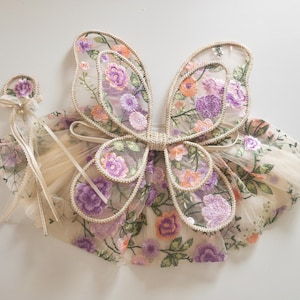 May include: A set consisting of a pair of sheer fairy wings and a matching wand, both crafted from beige mesh fabric featuring an intricate embroidered floral pattern in shades of purple, orange, and green with a decorative cream ribbon trim.