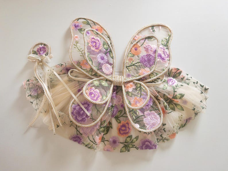 May include: A set consisting of a pair of sheer fairy wings and a matching wand, both crafted from beige mesh fabric featuring an intricate embroidered floral pattern in shades of purple, orange, and green with a decorative cream ribbon trim.