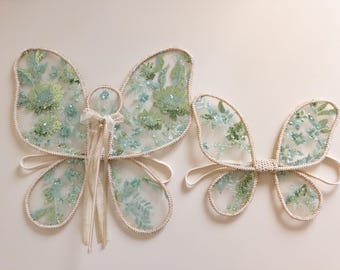 Puede incluir: Dos juegos de alas de mariposa hechas de tela de malla transparente con patrones florales bordados en verde y azul claro. Las alas están delineadas con un borde de color claro e incluyen correas elásticas, con el juego más grande incluyendo una varita central.