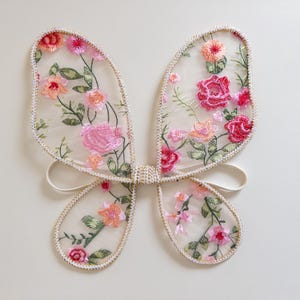 May include: Delicate butterfly wings made of sheer fabric, adorned with embroidered pink, peach, and red flowers and green leaves. The wings are outlined with a cream-colored trim and have white elastic straps for wear. A whimsical accessory.