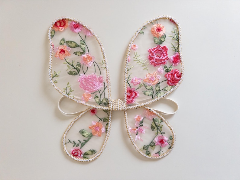 May include: Delicate butterfly wings made of sheer fabric, adorned with embroidered pink, peach, and red flowers and green leaves. The wings are outlined with a cream-colored trim and have white elastic straps for wear. A whimsical accessory.