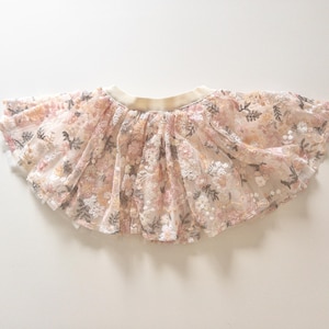 May include: A flared skirt featuring a sheer, layered tulle overlay with an intricate floral embroidery pattern in shades of soft pink, cream, and muted green. The garment includes a wide, solid cream-colored elastic waistband at the top.