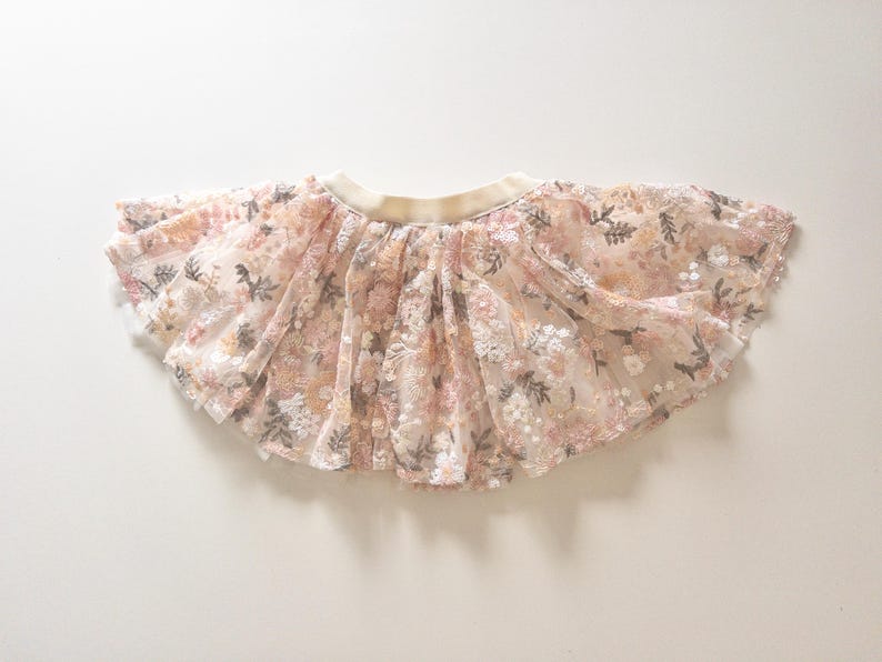 May include: A flared skirt featuring a sheer, layered tulle overlay with an intricate floral embroidery pattern in shades of soft pink, cream, and muted green. The garment includes a wide, solid cream-colored elastic waistband at the top.