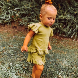 Op de afbeelding: Een jong kind draagt een groen geruit topje en een bijpassende short. Het kind heeft blond haar dat in een knotje is geknoopt met een bruine scrunchie. Het kind staat in een grasachtig gebied.