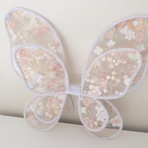 May include: A pair of butterfly wings crafted from sheer, light-colored fabric featuring an intricate floral embroidery pattern in shades of soft pink, white, and pale green, accented with shimmering sequins and finished with a white crocheted trim.