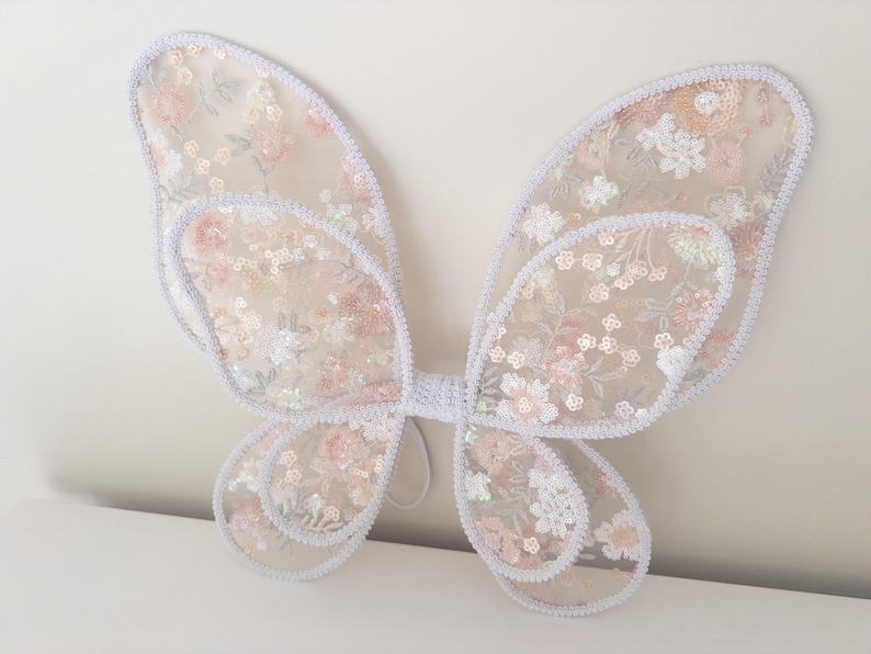 May include: A pair of butterfly wings crafted from sheer, light-colored fabric featuring an intricate floral embroidery pattern in shades of soft pink, white, and pale green, accented with shimmering sequins and finished with a white crocheted trim.