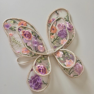 May include: A pair of butterfly wings made from sheer, translucent fabric featuring intricate floral embroidery in shades of purple, orange, and green. The edges of the wings are finished with a metallic gold trim, and a thin elastic strap is attached.