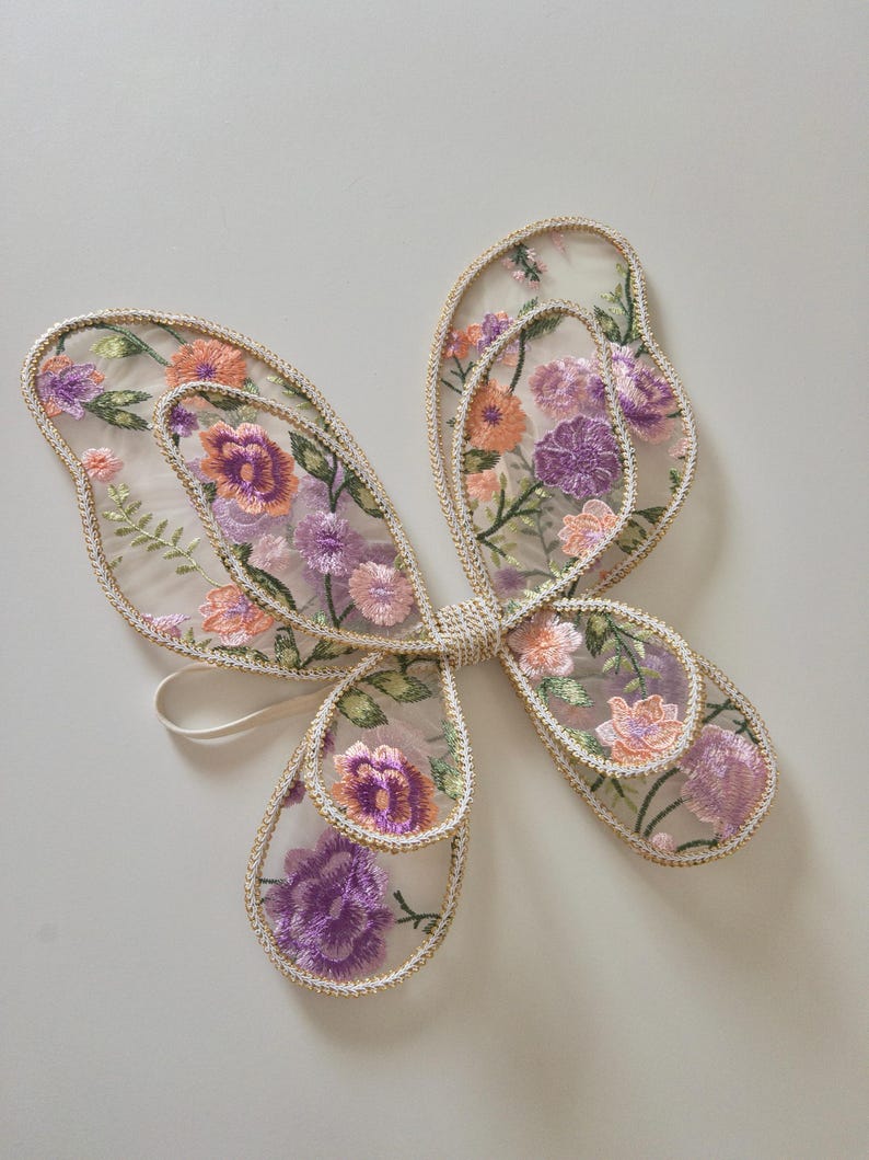 May include: A pair of butterfly wings made from sheer, translucent fabric featuring intricate floral embroidery in shades of purple, orange, and green. The edges of the wings are finished with a metallic gold trim, and a thin elastic strap is attached.