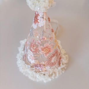 May include: A conical party hat made of sheer beige mesh fabric featuring embroidered floral patterns in shades of pink, peach, and white with shimmering sequins. The hat is topped with a fluffy white pom-pom and has a ruffled white lace trim.