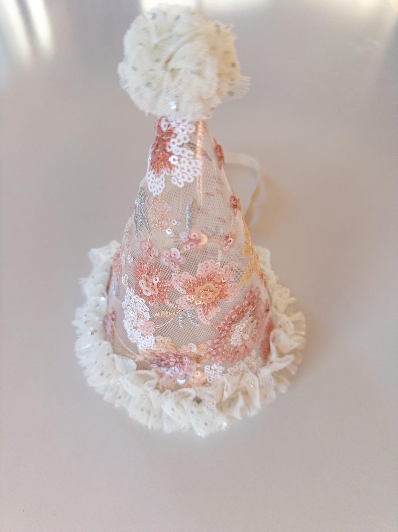 May include: A conical party hat made of sheer beige mesh fabric featuring embroidered floral patterns in shades of pink, peach, and white with shimmering sequins. The hat is topped with a fluffy white pom-pom and has a ruffled white lace trim.