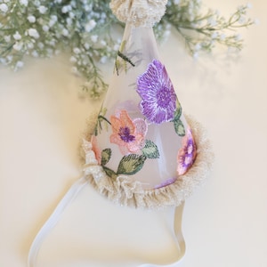 May include: A conical party hat made of sheer white fabric featuring embroidered purple and peach flowers with green leaves. The hat is topped with a textured beige pom-pom, finished with a ruffled beige trim at the base, and includes a white elastic strap.