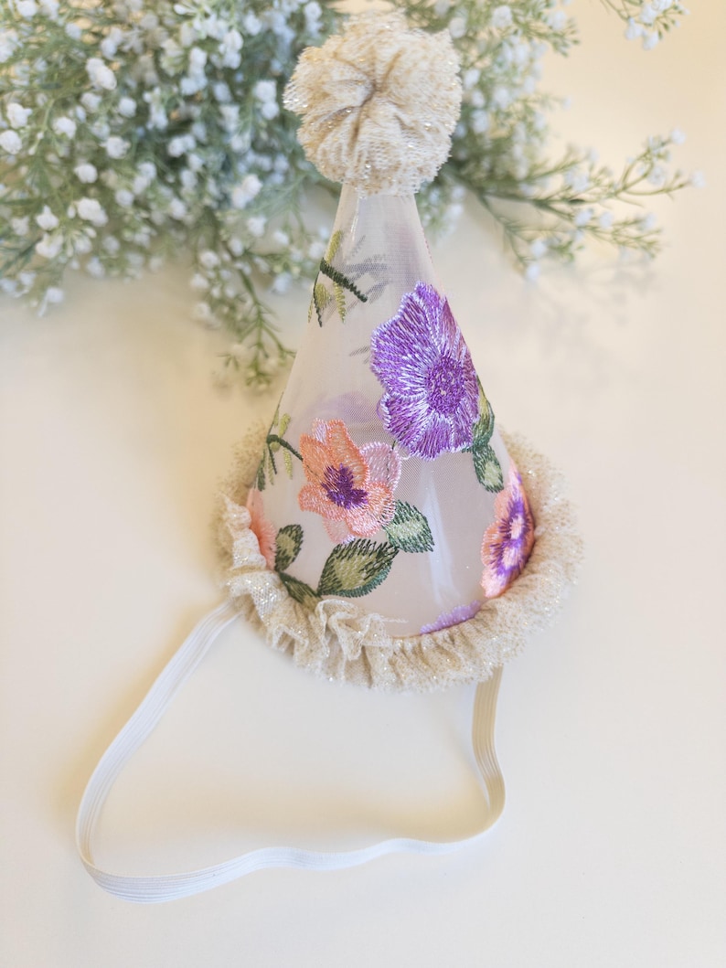 May include: A conical party hat made of sheer white fabric featuring embroidered purple and peach flowers with green leaves. The hat is topped with a textured beige pom-pom, finished with a ruffled beige trim at the base, and includes a white elastic strap.