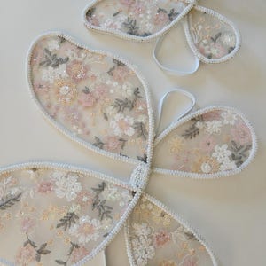 May include: Two sets of butterfly wings made from sheer, off-white mesh fabric. The wings feature intricate floral embroidery in shades of soft pink, cream, and sage green, accented with small, shimmering sequins and finished with white elastic straps.