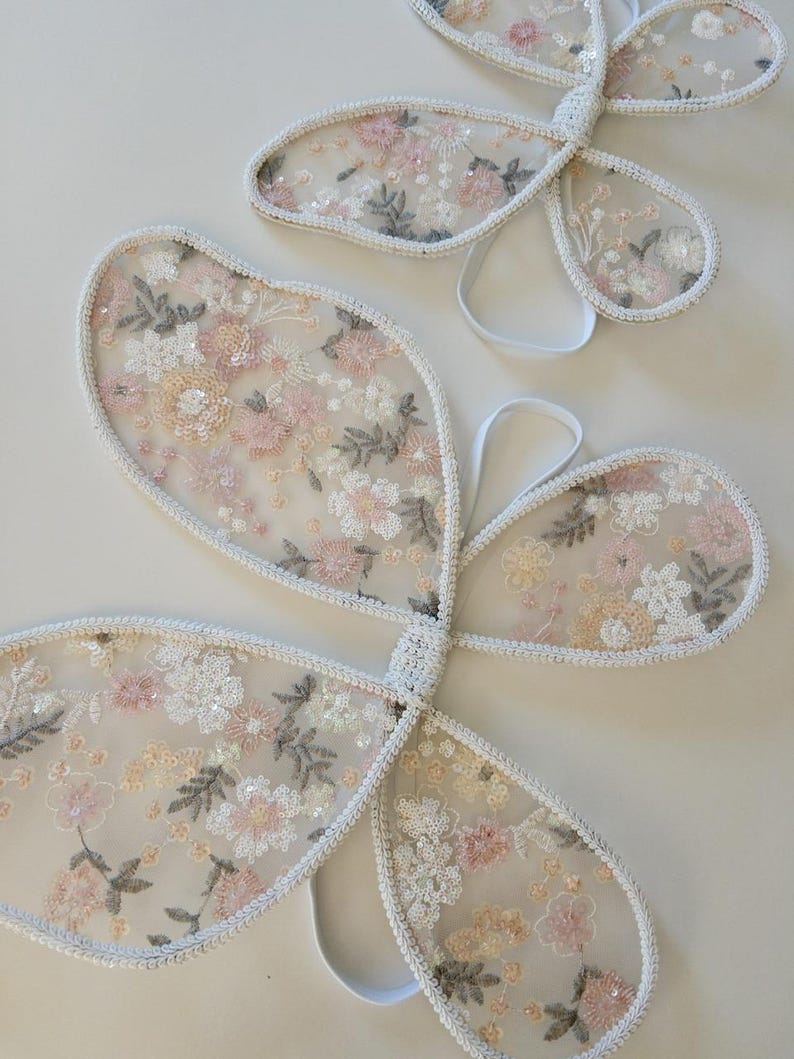 May include: Two sets of butterfly wings made from sheer, off-white mesh fabric. The wings feature intricate floral embroidery in shades of soft pink, cream, and sage green, accented with small, shimmering sequins and finished with white elastic straps.