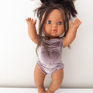 Puede incluir: Una muñeca con el pelo castaño en un moño desordenado que lleva un body de terciopelo morado. La muñeca tiene la piel morena y los ojos azules.