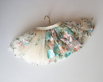 May include: A tulle skirt with floral embroidery in shades of pink, green, and blue. The skirt is cream colored and has a gold ribbon waistband.